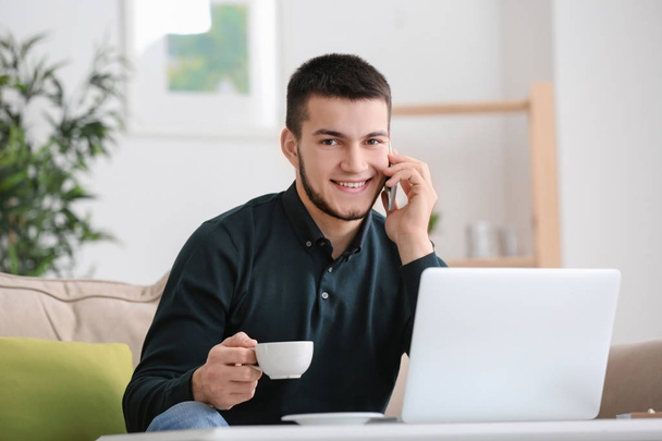 Young man using modern laptop and talking on mobile phone at home - Photo, image