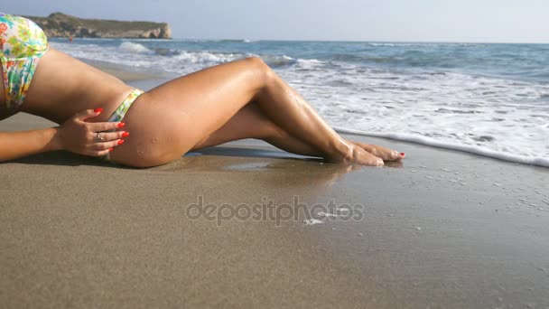 Wellen überspülen junge weibliche Füße. Eine nicht wiederzuerkennende attraktive Frau im Bikini liegt am Strand. Gebräuntes Mädchen sonnt sich an einem schönen Ufer. Sommerurlaub oder Urlaub. Nahaufnahme Zeitlupe - Filmmaterial, Video