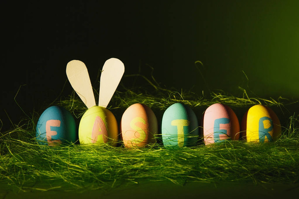Ряд из шести красочных пастельных монофонических окрашенных пасхальных яиц с надписью Пасха, веселые кроличьи уши на яйце в зеленой траве изолированы на темно-зеленом фоне с тенями и светом. Для рекламы
. - Фото, изображение
