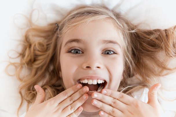 Draufsicht des lächelnden entzückenden Kindes, das auf dem Bett liegt und den Mund mit den Händen bedeckt - Foto, Bild