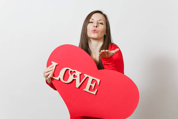 Young woman in red casual clothes sends air kiss, holds big red heart, wooden word love on white background. Copy space for advertisement. St. Valentine's Day or International Women's Day concept. - Zdjęcie, obraz