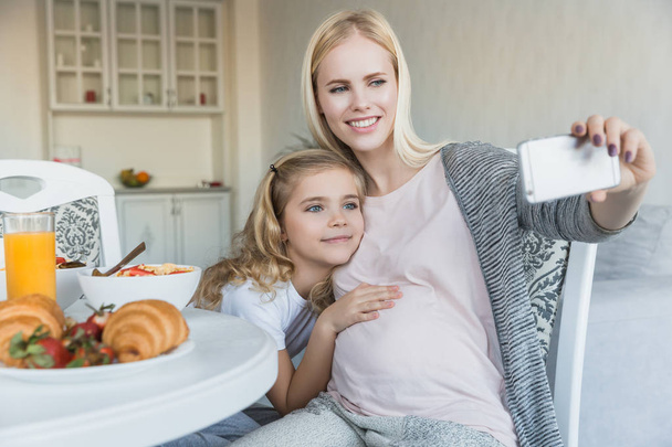 smiling pregnant mother and daughter taking selfie in kitchen - Foto, imagen