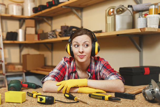 Pretty caucasian young brown-hair woman in plaid shirt, gray T-shirt, noise insulated headphones, yellow gloves working in carpentry workshop at wooden table place with piece of wood, different tools. - Photo, Image