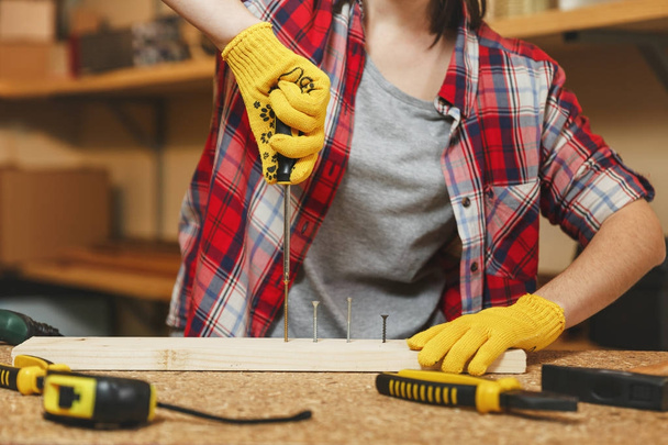 Close up of caucasian young woman in plaid shirt, gray T-shirt, yellow gloves twisting by screwdriver screw, working in carpentry workshop at wooden table place with piece of wood, different tools. - Photo, Image