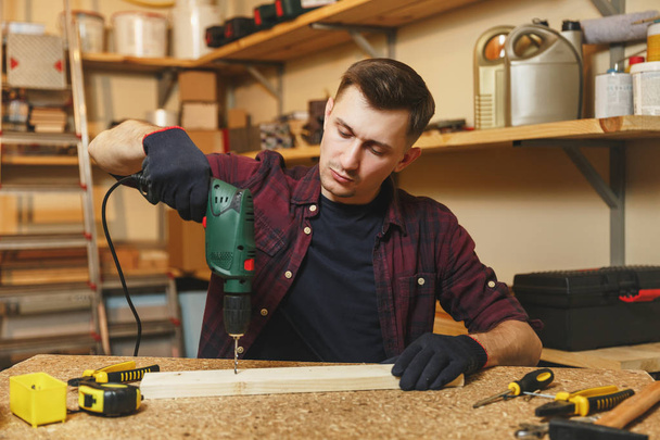 Handsome smiling caucasian young man in plaid shirt, black T-shirt, gloves drilling with power drill in piece of wood, working in carpentry workshop at wooden table place with different tools. - Photo, Image