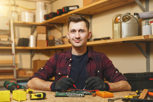 Multimeter for electrician. Caucasian young man in plaid shirt, black T-shirt digital electronic engineer repairing, soldering computer PC motherboard in workshop at wooden table with different tools. - Photo, Image
