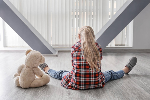 Little girl with teddy bear sitting on floor in empty room. Autism concept - Фото, изображение