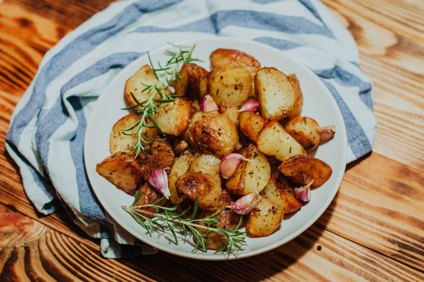 Roasted potato in white plate on wooden background with rosemary and garlic - Zdjęcie, obraz