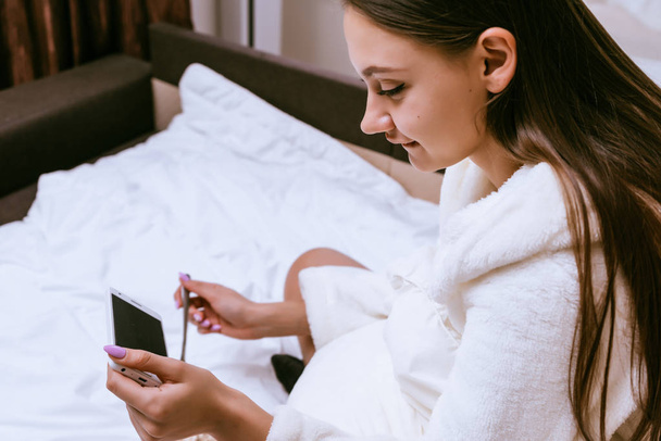 a woman in a bathrobe sits on a bed and has breakfast and looks into the phone - Photo, image