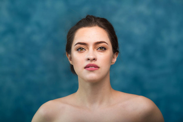 Young attractive girl with black hair fixed behind, big eyes, thick eyebrows and naked shoulders holding hand near face at gray background, portrait, copy space. - Photo, image