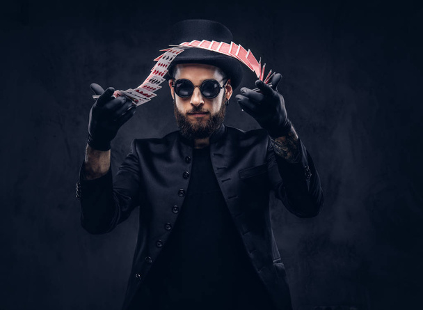 Magicien en costume noir, lunettes de soleil et chapeau haut de forme, montrant astuce avec des cartes à jouer sur un fond sombre
. - Photo, image