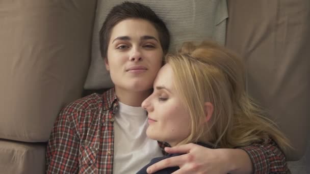 Two young lesbian girls lie on the couch, hug, cuddle, sleep, girl with short hair looks at the camera, lgbt family concept, happy, top shot 60 fps - Video