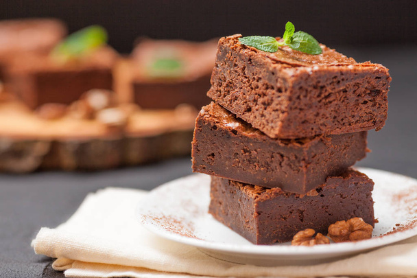 Chocolate brownie square pieces in stack on white plate with walnuts, decorated with mint leaves and cocoa on black background. Delicious dessert. Dark mood. Close up photography. Selective focus - Foto, immagini