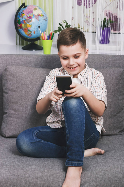 lächelnder Junge mit Telefon sitzt auf dem Sofa, weicher Hintergrund mit Fokus - Foto, Bild