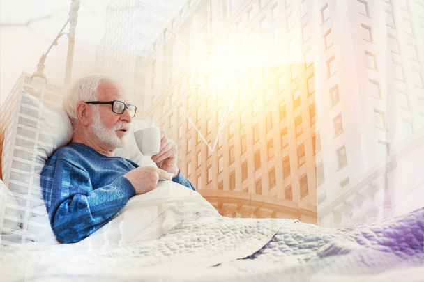 Aged man staying in bed and drinking hot coffee - Foto, afbeelding