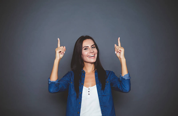 Lachende aantrekkelijke jonge vrouw in jeans overhemd punten met vingers over zichzelf in een reclame-zone op een grijze achtergrond, geïsoleerd. - Foto, afbeelding