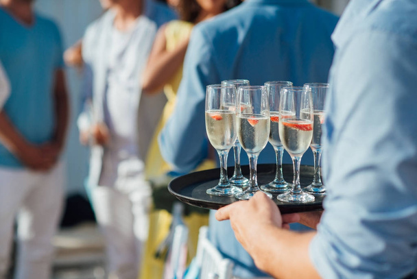 El camarero lleva una bandeja con copas de champán frío con fresas en el fondo de los invitados en trajes azules
 - Foto, Imagen