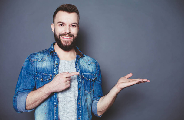 How do you like this product? A handsome smiling modern man in a denim shirt points a finger at his palm against a gray background. - Fotoğraf, Görsel