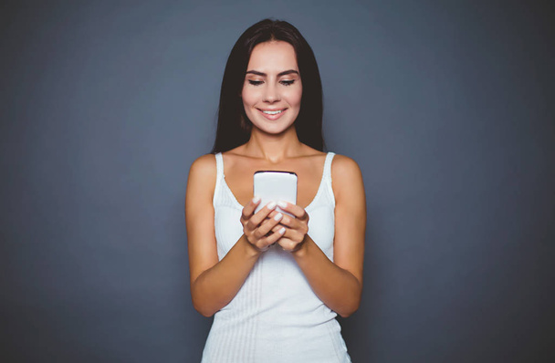 Wie geht es Ihnen? lächelnde attraktive moderne Frau hält ein Telefon in der Hand und schreibt eine Nachricht an ihren geliebten Mann vor grauem Hintergrund isoliert. - Foto, Bild