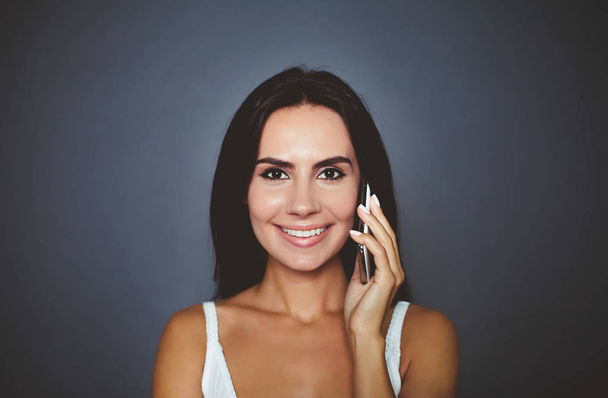 Communicate with your friends. Portrait of a beautiful smiling young woman who speaks by phone and looks at the camera on a gray background isolated. - Fotoğraf, Görsel