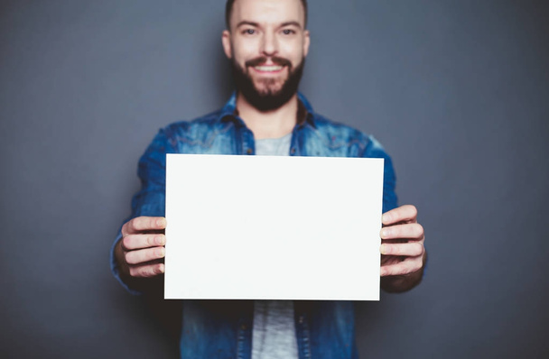 Everything is for you. Handsome young smiling man in a denim shirt shows a white sheet of paper in the camera on a gray background. Area for advertising. - Foto, Bild