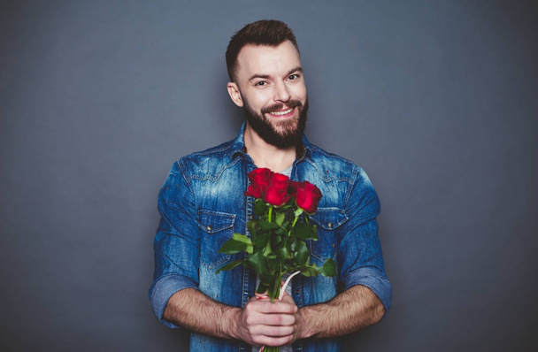 Cadeau voor de geliefde. Mooie glimlachend bebaarde man in een denim shirt heeft dat een rode roos tegen een grijze achtergrond. - Foto, afbeelding