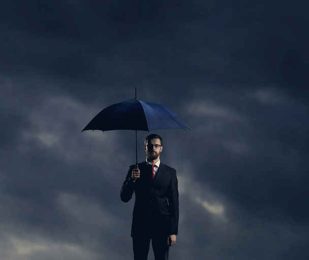homme d'affaires en costume noir tenant un parapluie et debout contre un ciel nuageux orageux. Entreprise, faillite, crise, concept d'assurance
 - Photo, image