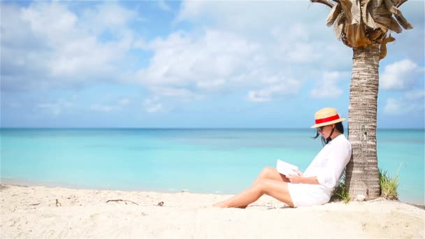 Mladá žena čte knihu na bílé pláži sedí pod palmového - Záběry, video