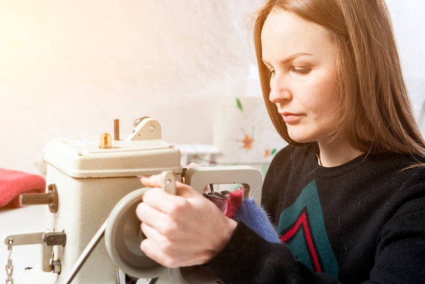Una mujer joven centelleante, peletero mira de cerca y cose una capa de piel brillante de piel azul natural en la máquina de coser
 - Foto, Imagen