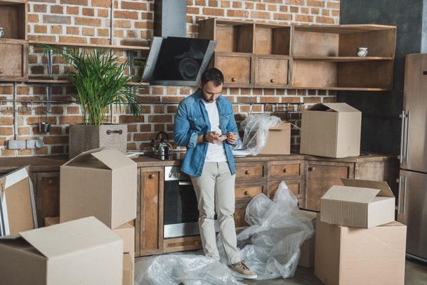 man using smartphone while standing between cardboard boxes in new apartment - Foto, Imagen