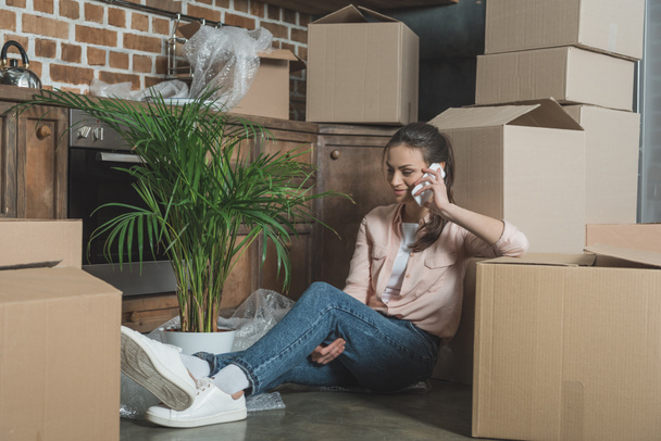 smiling young woman talking by smartphone while sitting between cardboard boxes in new apartment - Foto, Bild