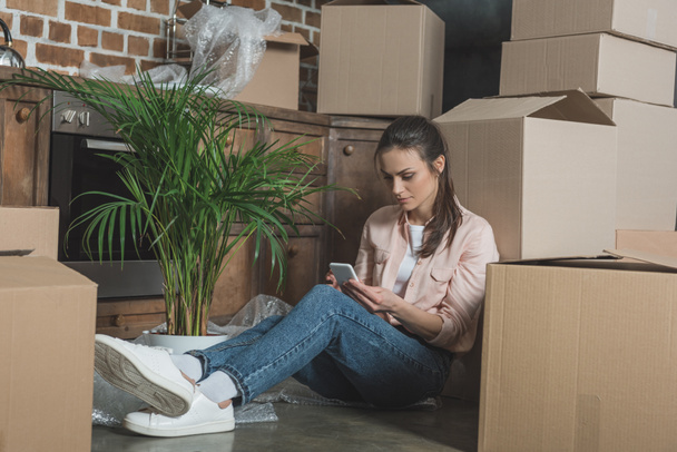 young woman using smartphone while sitting between cardboard boxes in new apartment - Photo, Image