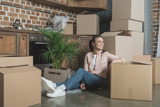 feliz joven mujer sonriendo y mirando hacia otro lado mientras se sienta con cajas de cartón en nuevo apartamento
 - Foto, Imagen