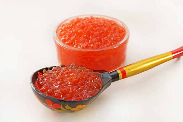 Caviar rojo en un tazón sobre fondo blanco. Cuchara de madera pintada en estilo tradicional Khokhloma con caviar natural. souvenir tradicional de Rusia
. - Foto, Imagen