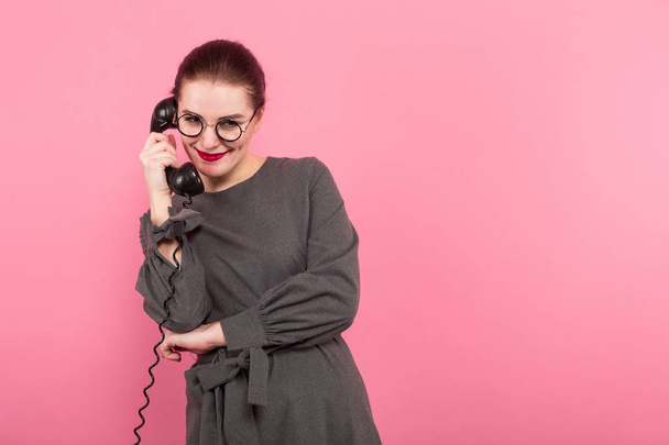 Attractive young brunette woman in eyeglasses talking on old phone against pink background  - Foto, Imagen