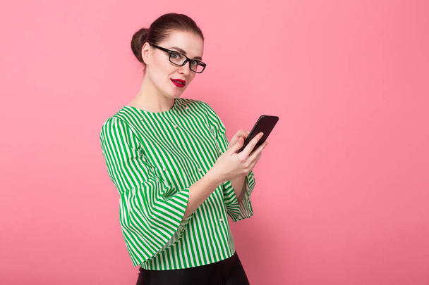 Kaunis nuori nainen tyylikäs silmälasit älypuhelimella vaaleanpunainen studio tausta
  - Valokuva, kuva
