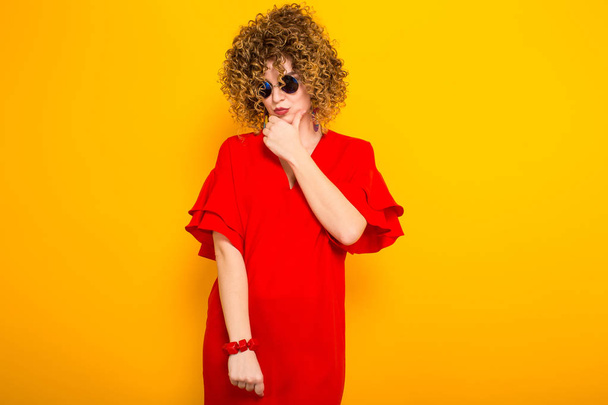 Attraente giovane donna dai capelli ricci in abito rosso e occhiali da sole pensando
 - Foto, immagini