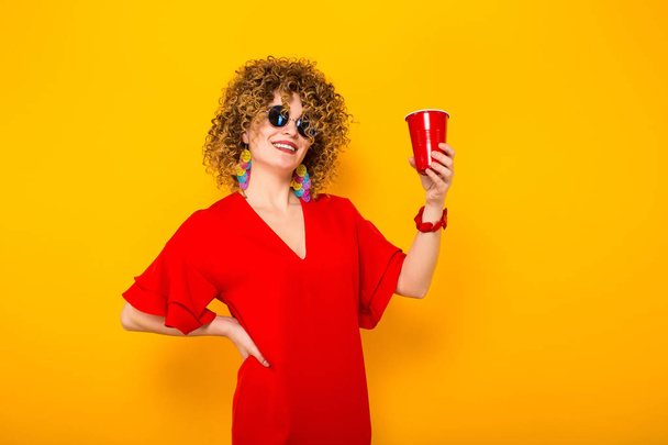 Çekici genç kıvırcık saçlı kadın kırmızı elbise ve plastik bardak içki ile tutarak güneş gözlüğü  - Fotoğraf, Görsel