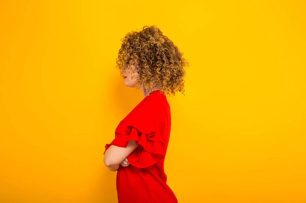 Boční pohled atraktivní kudrnaté vlasy dívka v červených šatech a sluneční brýle na žlutém podkladu - Fotografie, Obrázek