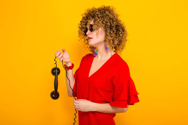 Attractive young curly haired woman in red dress and sunglasses holding handle of vintage phone - Foto, Imagem