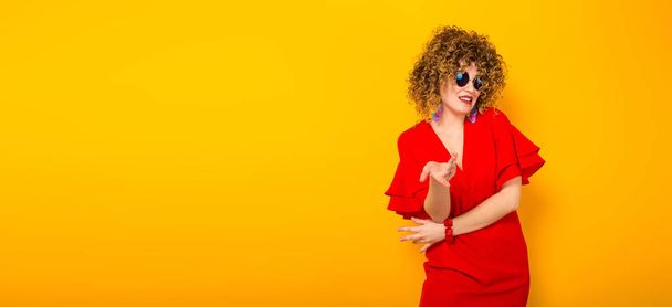 Attractive young curly haired woman in red dress and sunglasses pointing at camera - Foto, afbeelding