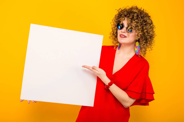 Привлекательная молодая кудрявая женщина в красном платье и солнцезащитных очках держит чистую карточку
 - Фото, изображение
