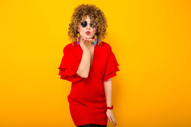 Attractive young curly haired woman in red dress and sunglasses blowing kiss - Photo, image