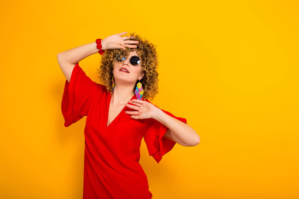 Attractive young curly haired woman in red dress and sunglasses posing against yellow background  - Foto, imagen