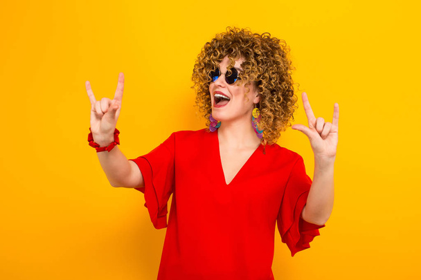 Atraktivní mladá žena kudrnaté vlasy v červených šatech a sluneční brýle zobrazující rock gesto - Fotografie, Obrázek
