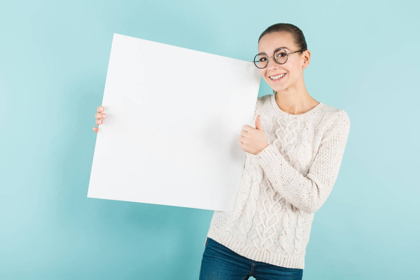 Pretty girl in stylish eyeglasses holding blank banner and showing thumb up  against blue studio background  - Foto, Imagen