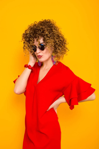 Attractive young curly haired woman in red dress and sunglasses posing against yellow background  - Valokuva, kuva
