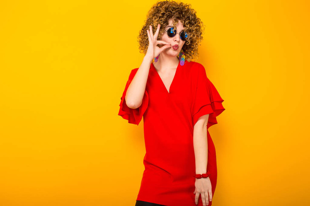 Attraente giovane donna dai capelli ricci in abito rosso e occhiali da sole mostrando gesto ok
  - Foto, immagini