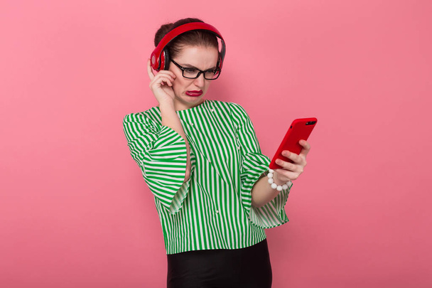 niedliche junge brünette Frau mit roten Kopfhörern hält Smartphone und macht lustiges Gesicht vor rosa Hintergrund - Foto, Bild