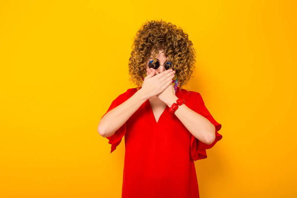 Attraente giovane donna dai capelli ricci in abito rosso e occhiali da sole che si copre la bocca con le mani
 - Foto, immagini